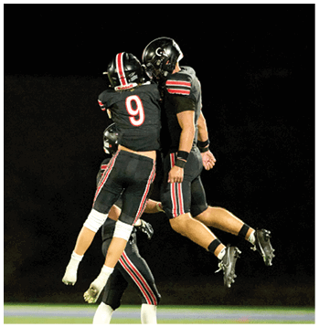 Two people wearing black football uniforms with red and white stripes and helmets are interacting closely on a dark field. Description generated by AI
