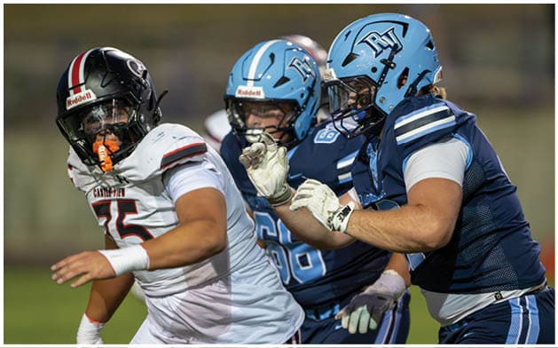 Two people in blue football uniforms and helmets are closely pursuing a person in a white football uniform and helmet during a game. Description generated by AI
