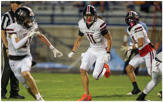 Three people in football uniforms are actively running on a grassy field during a game, with one person in the center wearing bright orange cleats. Description generated by AI
