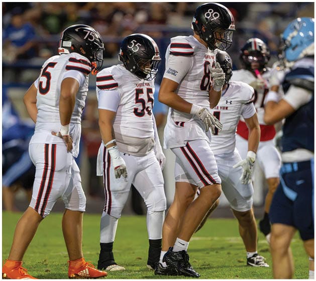 Four people in white football uniforms with black helmets stand on a grassy field during a game, with one person in a blue uniform nearby. Description generated by AI