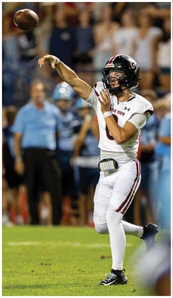 A person in a white football uniform is throwing a football on a grassy field with blurred people and a referee in the background. Description generated by AI