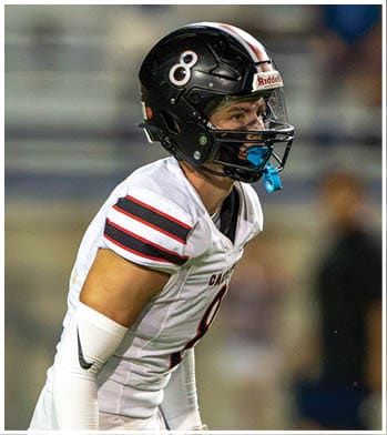 An adult wearing a white football uniform with black and red stripes, a black helmet, and gloves is standing on a field in a ready position. Description generated by AI