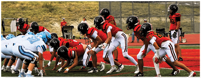 Two opposing American football teams in red and white and light blue and white uniforms are lined up facing each other on a field with a red track and grassy hill in the background. Description generated by AI