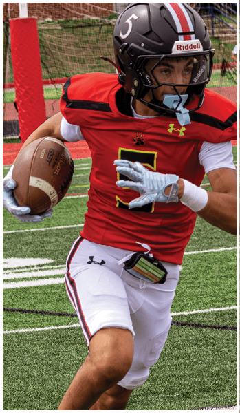 A person wearing a red and white football uniform with a black helmet is running on a field while holding a football. Description generated by AI