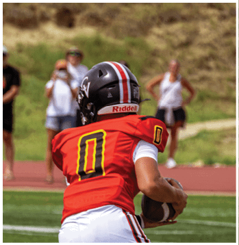 A person wearing a red football jersey with the number zero and a black helmet is holding a football on a field with blurred spectators in the background. Description generated by AI