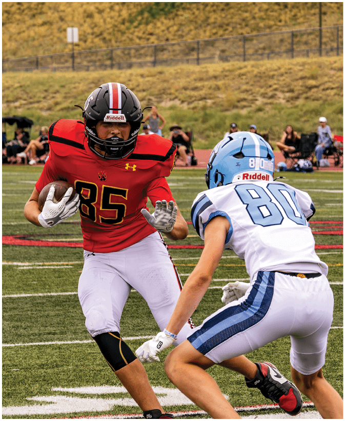 Two people in football uniforms are on a field with one person in a red jersey running with the ball while the other in a white and blue jersey prepares to block or tackle. Description generated by AI