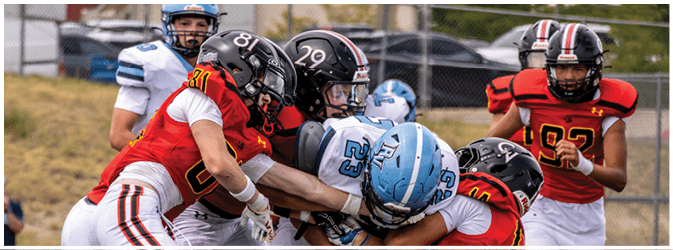Several people wearing football helmets and uniforms are engaged in a tackle on a grassy field with a chain-link fence and parked cars in the background. Description generated by AI