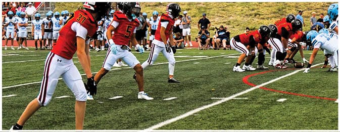 People in football uniforms are lined up on a field preparing for a play with a team in red jerseys facing a team in light blue jerseys. Description generated by AI