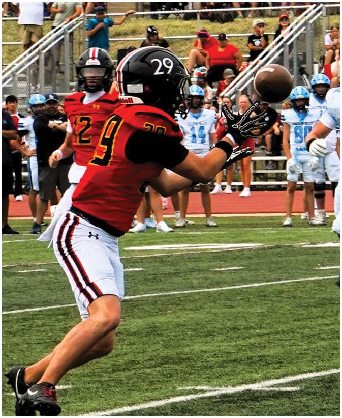A person in a red and black football uniform is reaching out to catch a football while players in white and blue uniforms approach on a grassy field with spectators in the background. Description generated by AI
