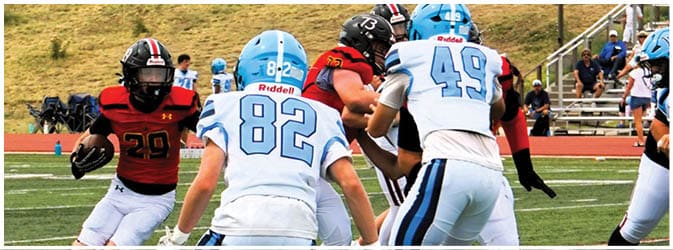 People in football uniforms are actively playing on a field with one person holding the ball and others preparing to block or tackle. Description generated by AI