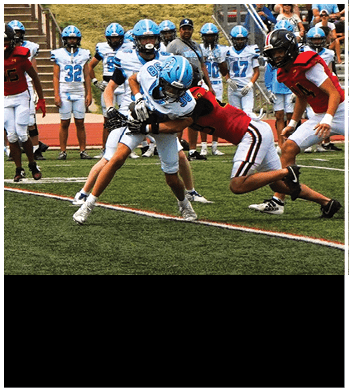 A person in a red sports uniform is tackling a person in a white and blue football uniform on a field while other players and a coach watch from the background. Description generated by AI