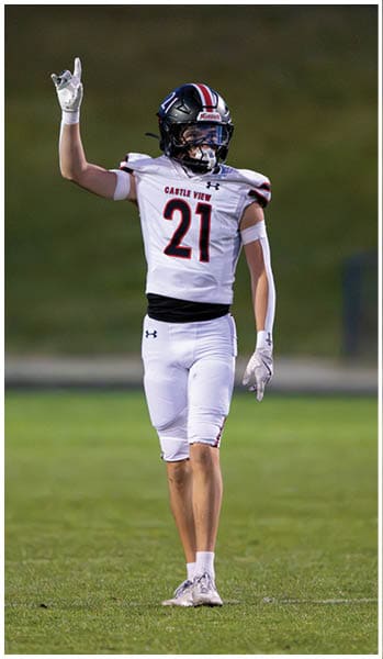 A person wearing a football uniform with the number 21 is standing on a grassy field with one arm raised and pointing upward. Description generated by AI