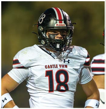 A person wearing a white football jersey with the number 18 and a black helmet stands on a field with a blurred background. Description generated by AI