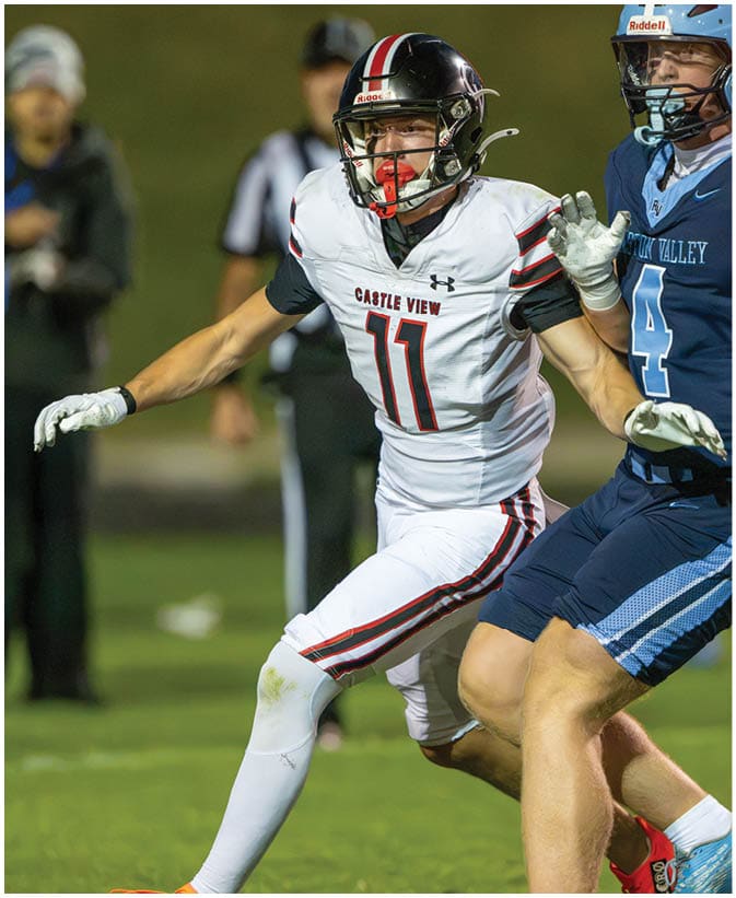 A person wearing a white football uniform with the number 11 and a black helmet is actively reaching out during a game with a blurred referee in the background. Description generated by AI