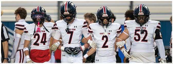 Four people in football uniforms stand arm in arm on a field, appearing focused and united before a game. Description generated by AI