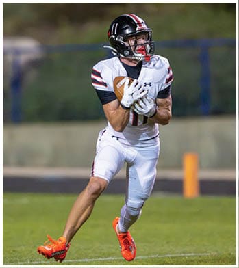 A person wearing a white football uniform with black and red stripes is running on a grassy field while holding a football securely. Description generated by AI