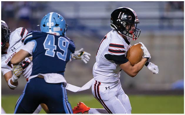 Two people in football uniforms are engaged in a play on a field, with one person holding the ball and the other preparing to tackle. Description generated by AI