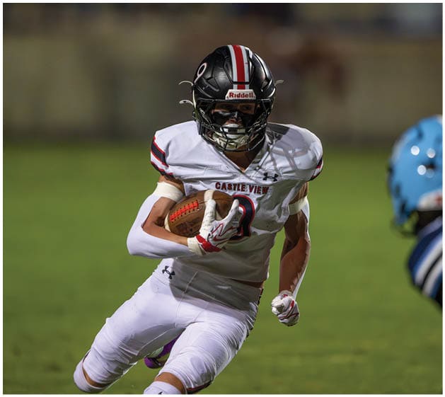 A person wearing a white football uniform with “Castle View" on the chest is running with a football while another person in a blue helmet and uniform approaches. Description generated by AI