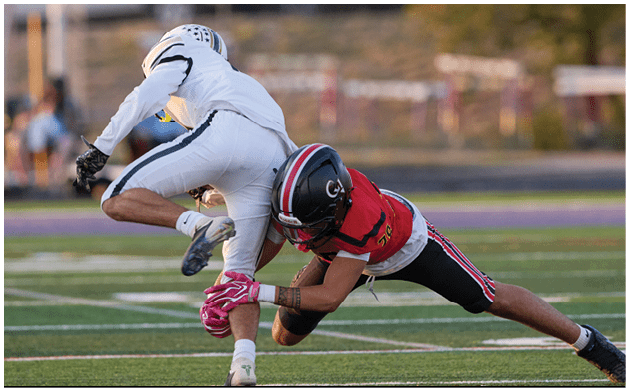 Two people in football uniforms are engaged in a tackle on a field with blurred vehicles and fencing in the background. Description generated by AI