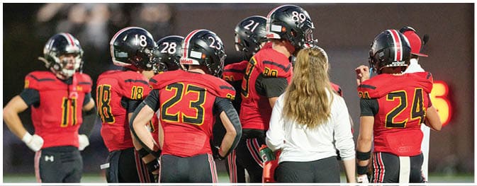 A group of people wearing red and black football uniforms are gathered on a field with one person in a white shirt standing among them. Description generated by AI