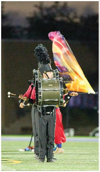 A person in a marching band uniform is playing a bass drum on a field while another person behind them waves a colorful flag. Description generated by AI