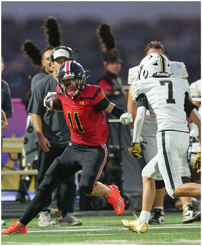 A person in a red and black football uniform is running while two people in white football uniforms with gold cleats are moving toward them on a football field. Description generated by AI