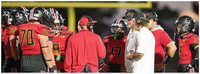 A group of people wearing football helmets and uniforms are gathered on a field with two people in headsets and casual clothing appearing to give instructions. Description generated by AI