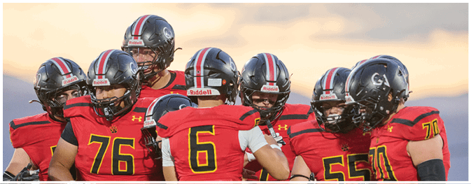 A group of people wearing red and black football uniforms and helmets are gathered closely together on a field during sunset. Description generated by AI