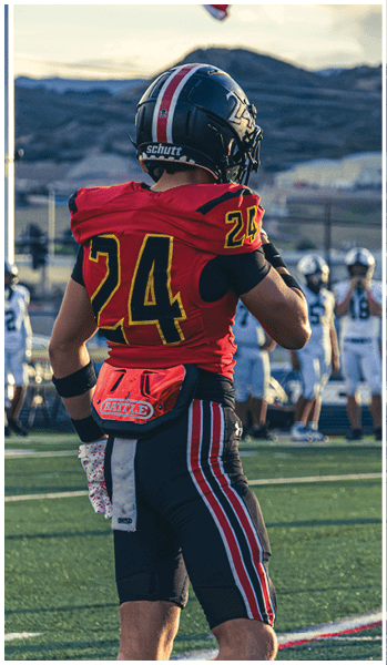 A person wearing a red and black football uniform with the number 24 and a black helmet is standing outdoors with a blurred mountainous background and a flag waving above. Description generated by AI