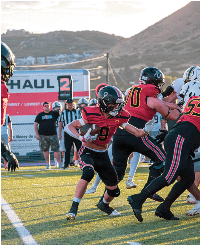 A group of people wearing football uniforms are actively playing on a field with one person holding the ball and others blocking or running. Description generated by AI