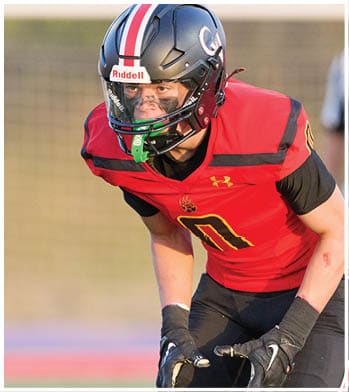 A person wearing a red and black football uniform and helmet is crouched in a ready position on a field. Description generated by AI