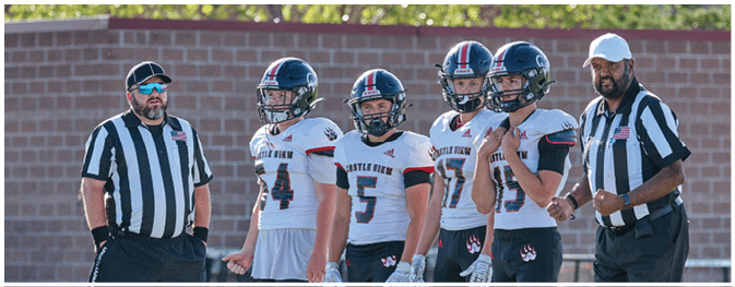 Five people wearing football uniforms and two referees in striped shirts stand together on a field with a brick wall and greenery in the background. Description generated by AI