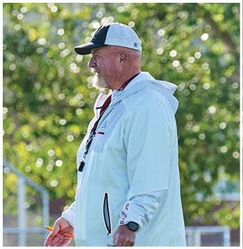 A person wearing a white hooded jacket and a baseball cap is standing outdoors with trees in the background, holding a whistle and a pen. Description generated by AI