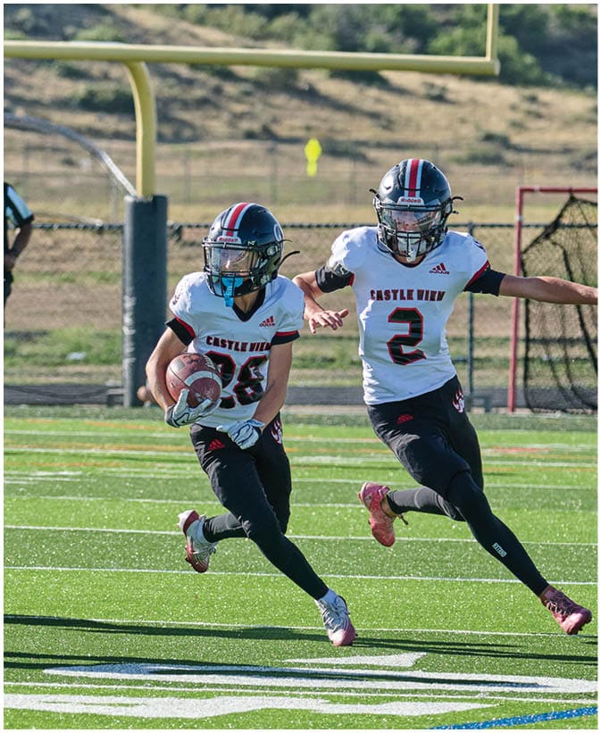 People in football uniforms are actively playing on a field with one person running with the ball while others block and chase, and a referee stands in the background near the goalpost. Description generated by AI