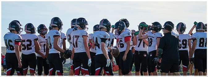 A group of people wearing football uniforms and helmets are gathered on a field with a coach giving instructions. Description generated by AI