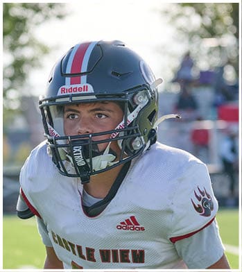 A person wearing a football helmet and white Adidas jersey with red and black details is on a field with blurred spectators and trees in the background. Description generated by AI