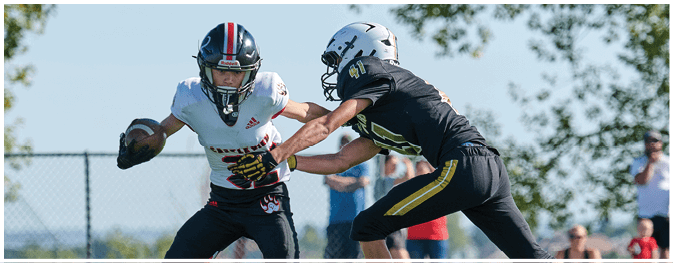 Two people wearing helmets and sports gear are playing football outdoors with trees and a clear sky in the background. Description generated by AI