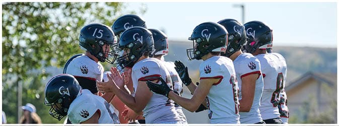 A group of people wearing white sports uniforms and black helmets are gathered closely outdoors, appearing to be engaged in a team discussion or huddle. Description generated by AI