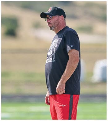 A person wearing a black cap, glasses, a black sports t-shirt, and red athletic pants is standing outdoors on a grassy field. Description generated by AI
