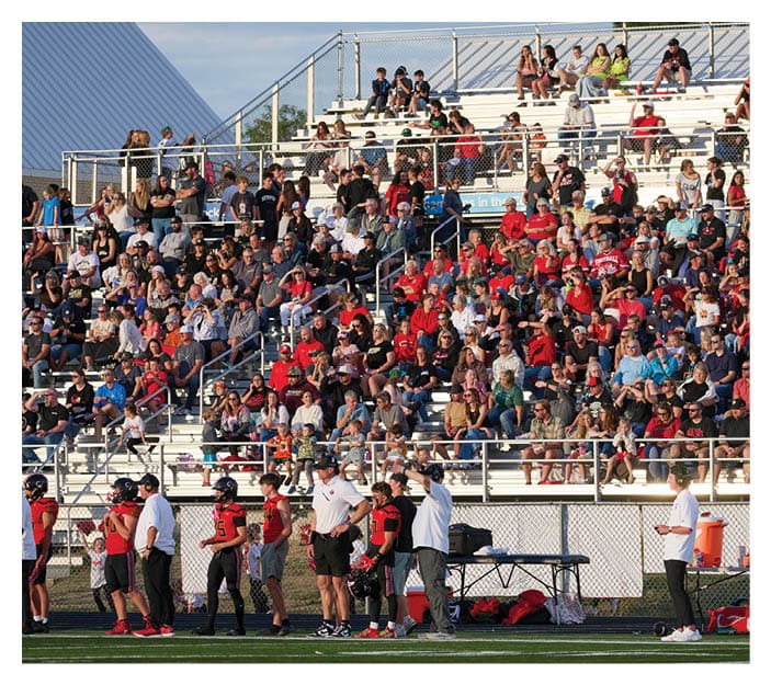 A large crowd of people is seated in bleachers watching a sports event while several players and coaches in red and black uniforms stand on the sidelines of a green field. Description generated by AI