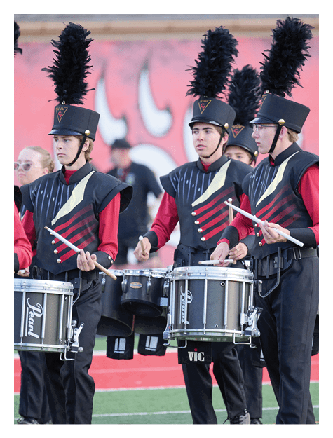 Three people wearing marching band uniforms with tall black feathered plumes on their hats stand together, with one holding drumsticks. Description generated by AI