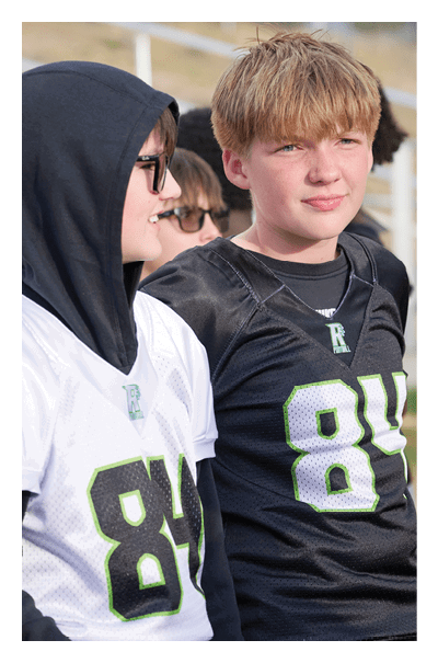 Two children wearing sports jerseys with the number 84 are standing outdoors near bleachers, one in a black jersey and the other in a white jersey with a hooded sweatshirt underneath. Description generated by AI