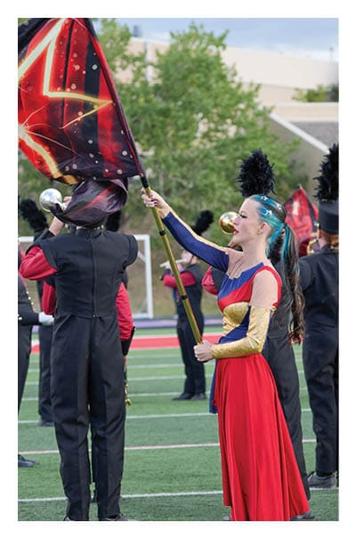 A person with long hair in a colorful red, blue, and gold costume is holding and waving a large flag with a star design while other people in black marching band uniforms with tall feathered hats stand on a sports field. Description generated by AI