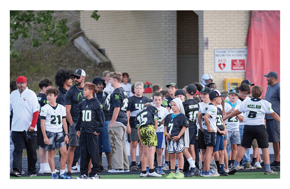A group of children wearing sports jerseys and caps are gathered together, some looking in different directions, with a brick wall and a sign in the background. Description generated by AI