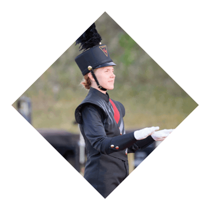 Person wearing a tall black marching band hat with a large black plume and a uniform, looking to the side outdoors. Description generated by AI