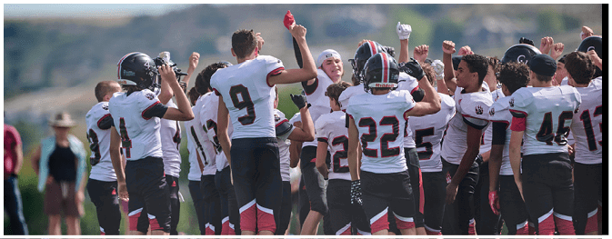 A group of people wearing sports uniforms and cleats stand on a grassy field with helmets placed on the ground nearby. Description generated by AI