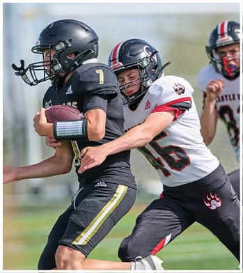 A person in a black football uniform is holding a football while being tackled by another person in a white and black football uniform on a grassy field with a third player running behind them. Description generated by AI