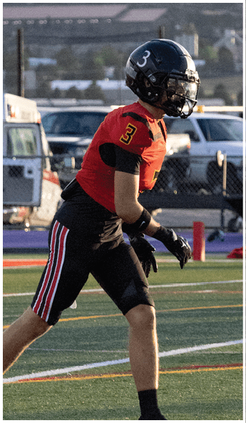A person wearing a black helmet and red sports jersey with the number 3 is standing on a football field preparing for action. Description generated by AI
