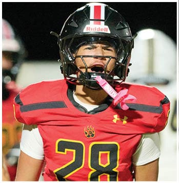 A person wearing a black football helmet and red jersey is shouting with a pink mouthguard hanging from the helmet. Description generated by AI