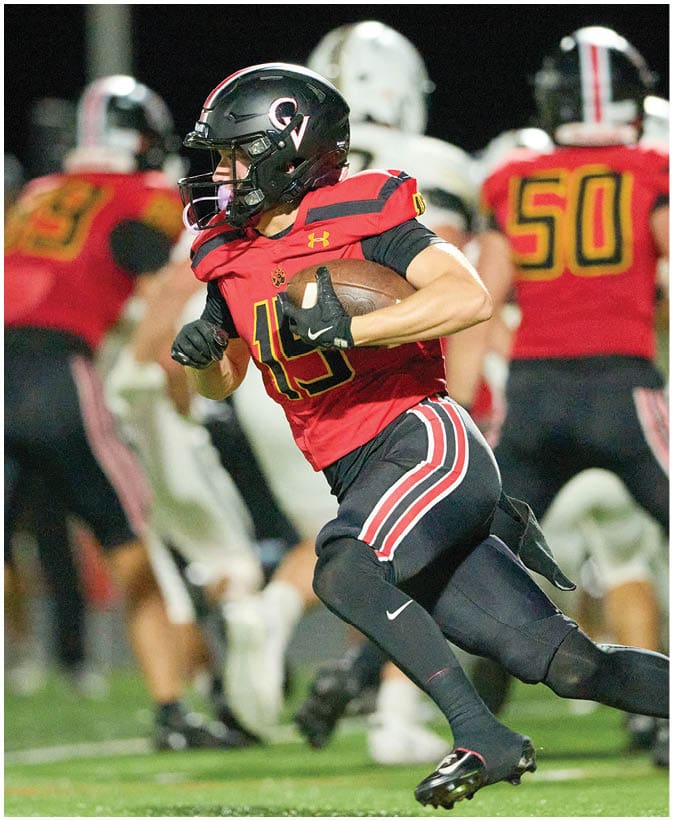 A person wearing a red and black football uniform is running with a football on a field during a game. Description generated by AI